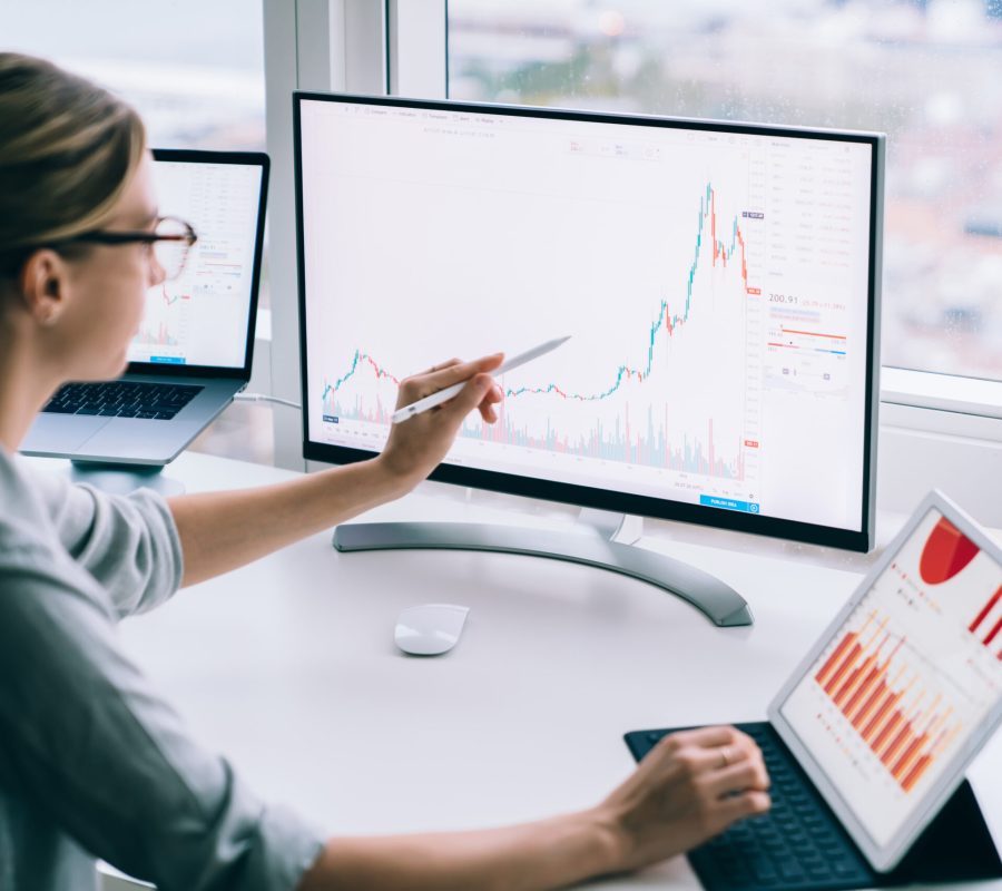 Side view of anonymous young female analyst pointing with stylus at desktop computer while studying chart near tablet at work