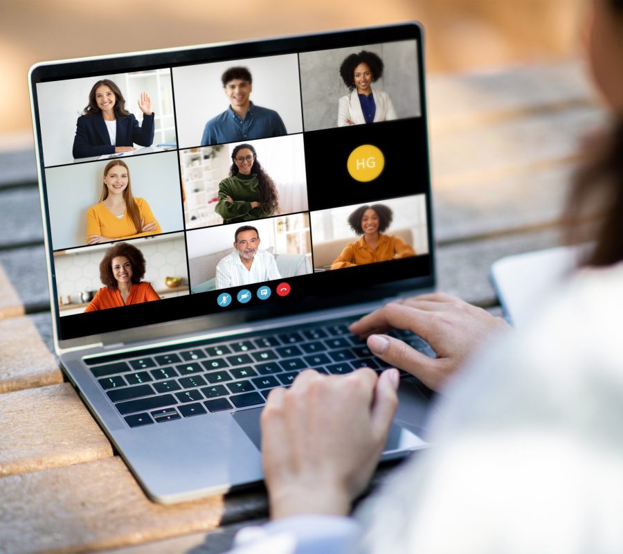 Unrecognizable woman sitting at a desk, typing on a laptop while video conferencing with a diverse group of people displayed on the screen. The individuals on screen are engaged in a virtual meeting